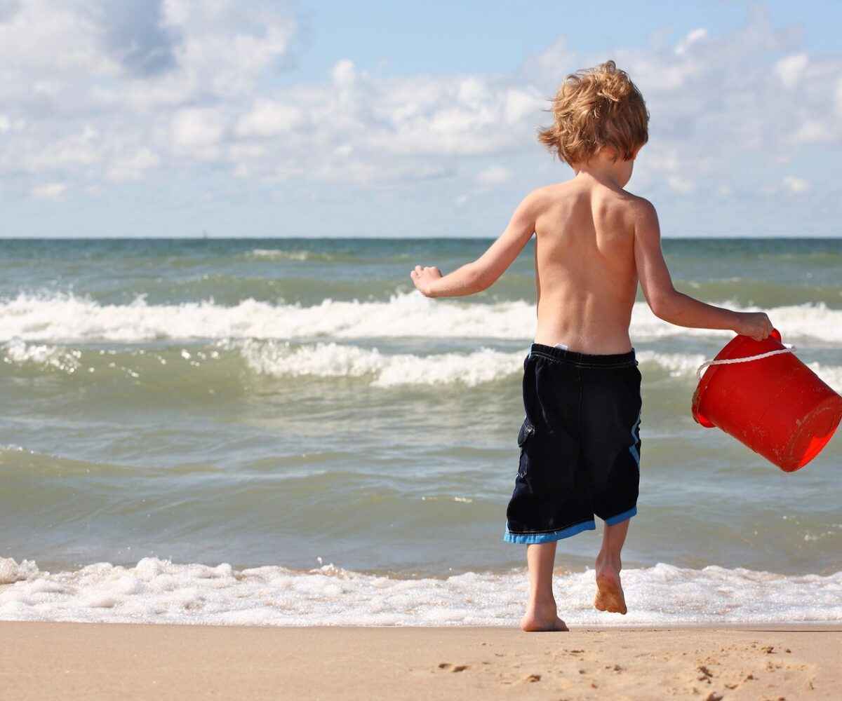 Little boy running into the ocean carrying a sand bucket.