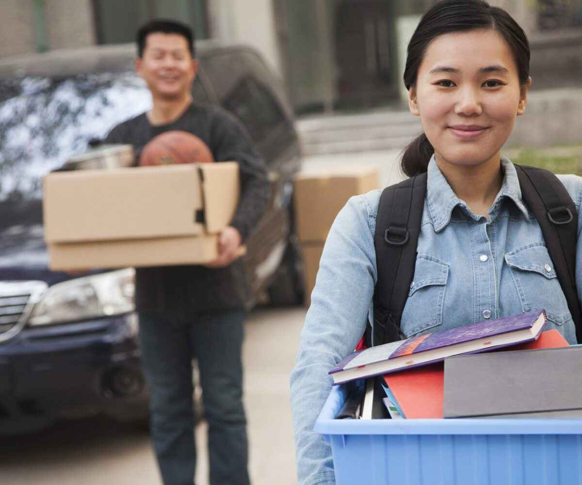 Asian college student carrying box of books with man in the background.