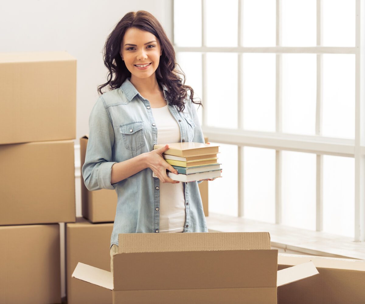 Young woman holding a stack of books surrounded by moving boxes.