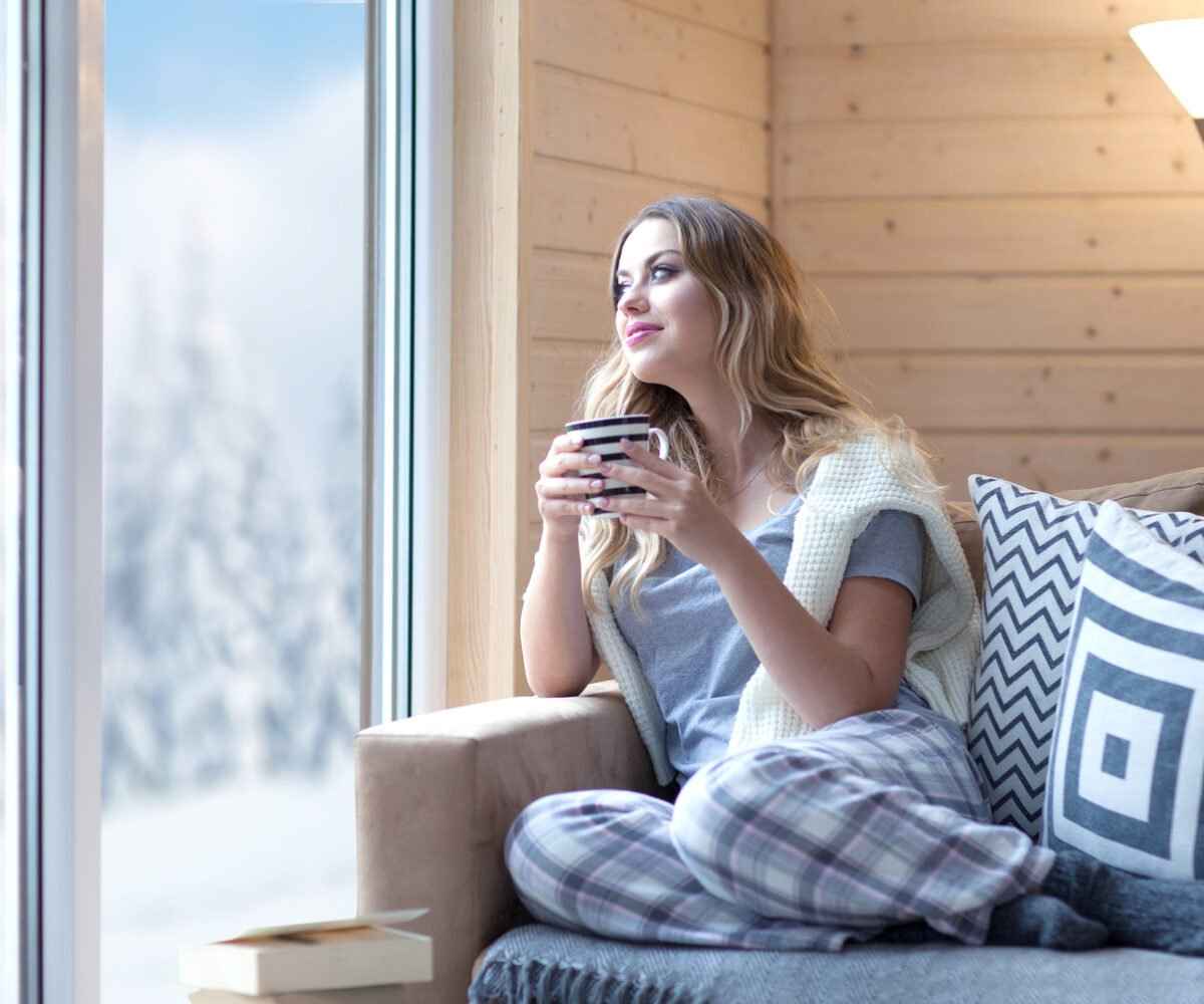 A young woman holding a mug looking out a window.