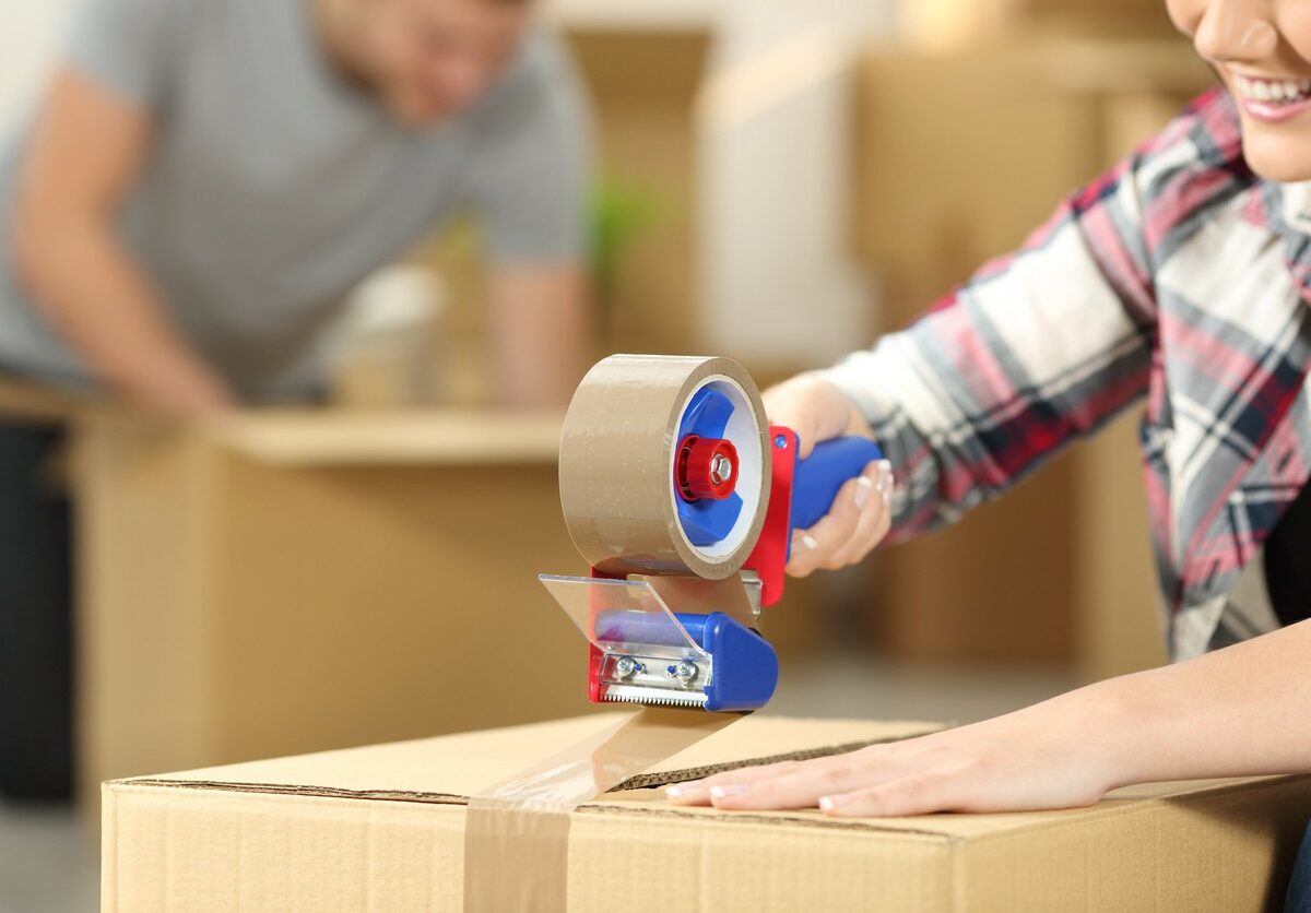 A woman taping the top of a box down with a roll of packing tape with their partner packing another box behind them.