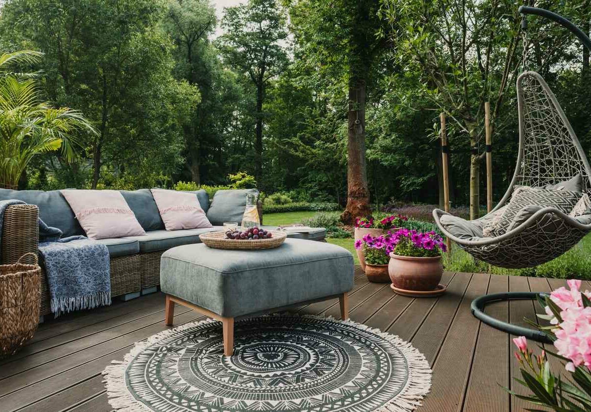 A patio garden is set up in a wooded area, with blue furniture and pink flowers set out on a stained deck.