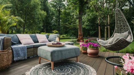 A patio garden is set up in a wooded area, with blue furniture and pink flowers set out on a stained deck.