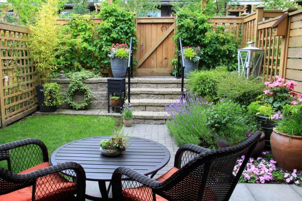 A patio garden spans out across a yard, with plants going up a flight of steps. Red chairs and a circular table are set up in the front of the garden.