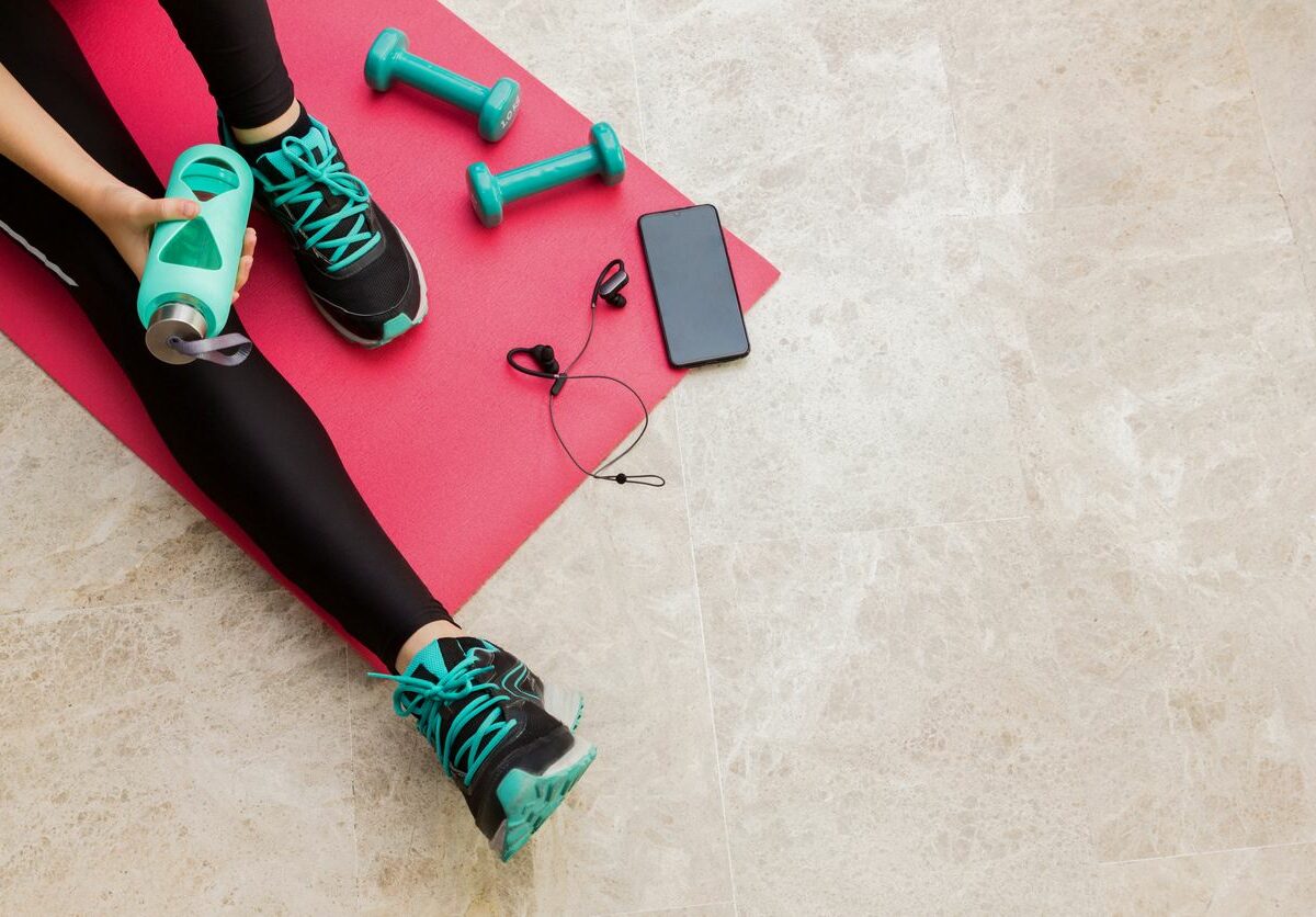 A woman in black exercise clothes sits on a pink yoga matt and holds a water bottle, surrounded by her cell phone, headphones, and blue dumbbells.