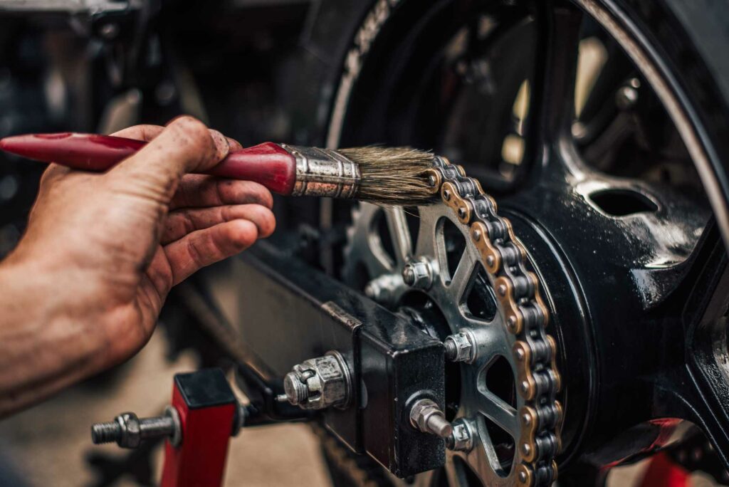 A man brushes lubrication onto their motorcycle’s chain using a red paint brush