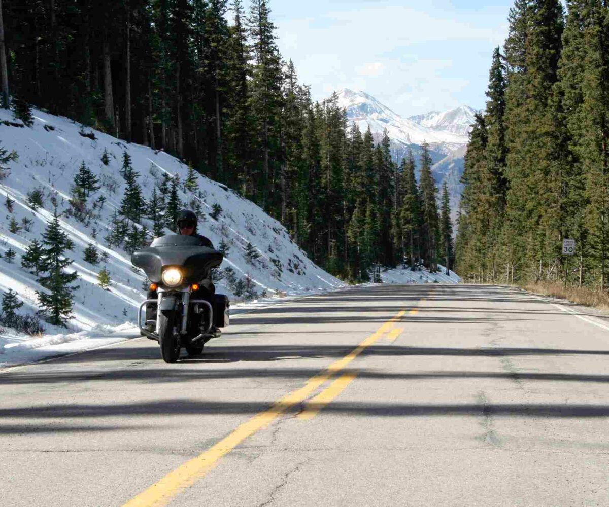 A rider drives their motorcycle through a mountain pass, with snow covering the ground and green pine trees surrounding them.