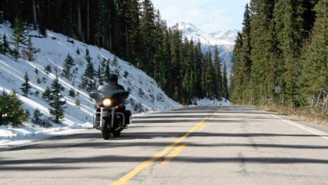 A rider drives their motorcycle through a mountain pass, with snow covering the ground and green pine trees surrounding them.