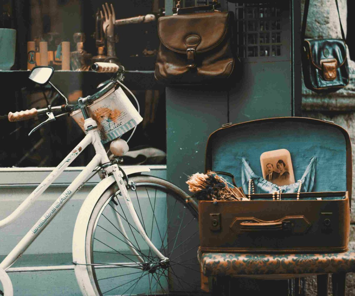 A vintage bicycle with a basket, a suitcase with a personal photo, dried flowers and jewelry along with personal bags hanging near a shop of antiques.
